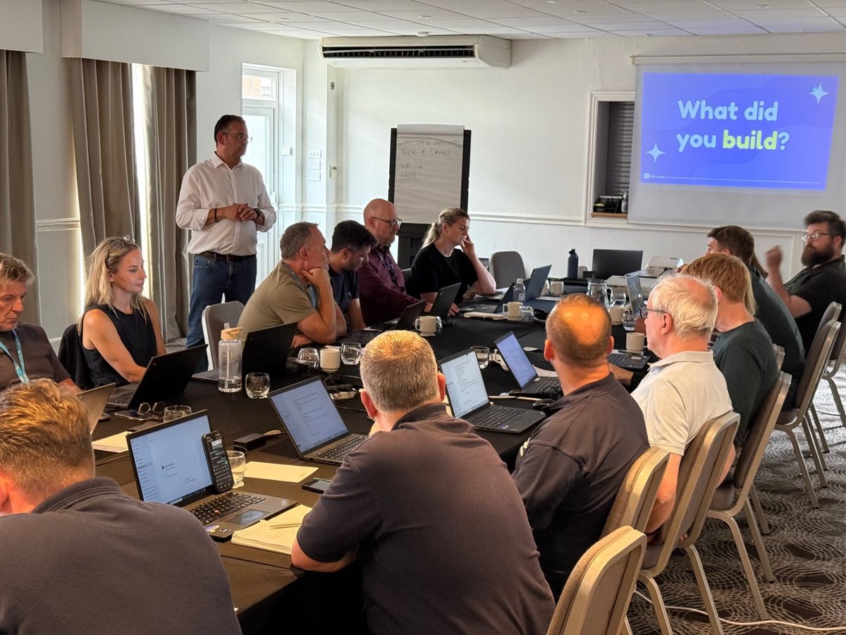 Group of professionals in business casual attire seated around tables with laptops during a workshop, with a presenter standing in the background and a screen displaying What did you build?